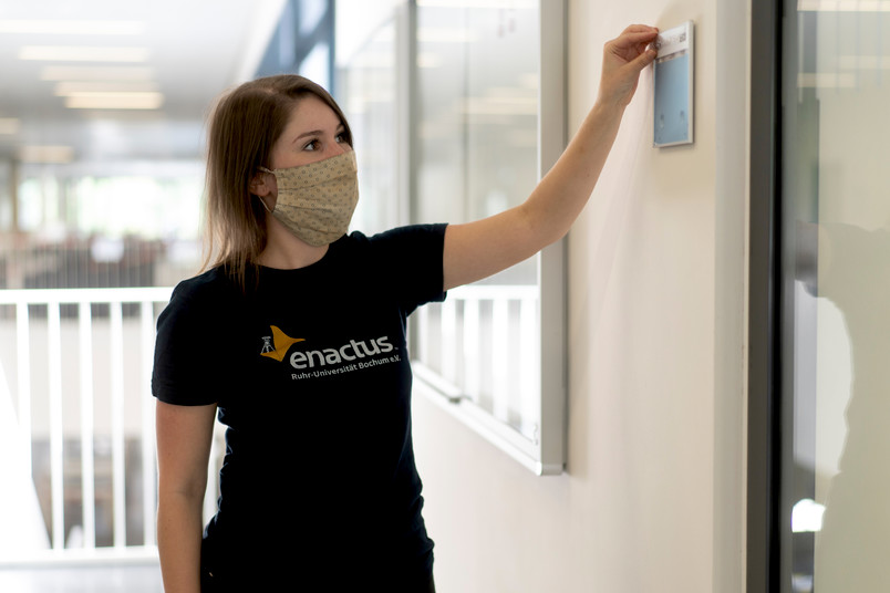 Eine junge Frau mit enactus RUB T-Shirt, die eine Braille Beschriftung an einem Raum der RUB anbringt.