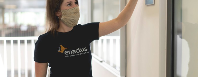Eine junge Frau mit enactus RUB T-Shirt, die eine Braille Beschriftung an einem Raum der RUB anbringt.