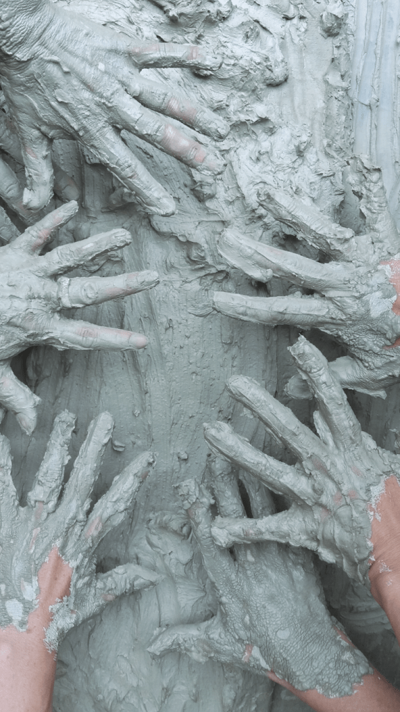 Es sind mehrere gespreizte Hände zu sehen, die sich in der Tischmitte treffen. Der Tisch und die Hände sind bedeckt mit hellem Ton.