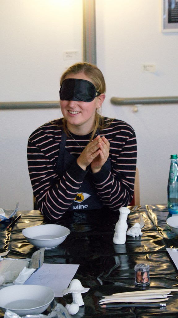 Eine Teilnehmerin befühlt ihren Ton mit verbundenen Augen. Um sie herum sind einige Materialien zu sehen. Außerdem einige fertige Tonkunstwerke: Eine kleine Flasche und ein Pilz. 