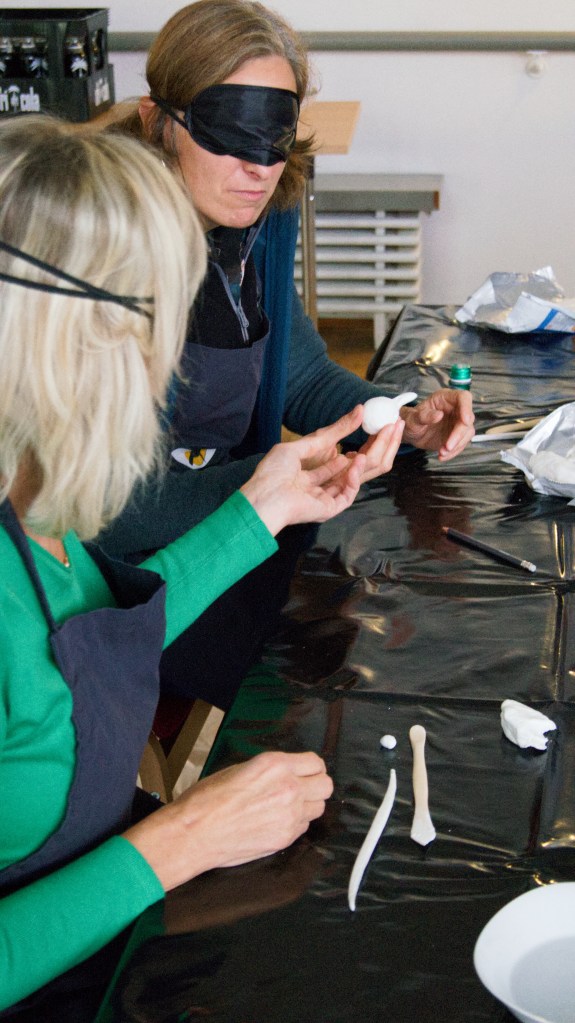 Zwei Workshopteilnehmerinnen sitzen am Tisch und reichen sich ihre Kunstwerke. Dabei haben sie schwarze Augenmasken auf.