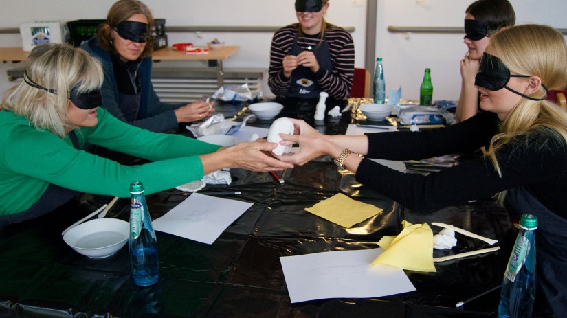 Mehrere Workshopteilnehmer sitzen mit verbundenen Augen am Tisch. Zwei von ihnen reichen sich über den Tisch hinweg einen Klumpen Ton. Die anderen Teilnehmer im Hintergrund formen ihre Figuren. 