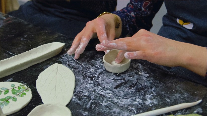 Eine Workshopteilnehmerin formt mit ihren Händen eine kleine Tonschale. Auf dem Tisch befinden sich daneben noch eine längliche Tonschale, ein Blatt aus Ton und ein Tonscheibe mit einem eingepressten Pflanzenzweig. 