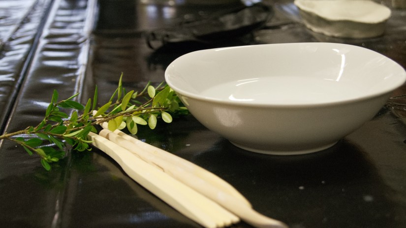 Einige unterer Materialien sind hier zu sehen. Tonwerkzeug aus Holz, eine weiße Wasserschale und im Hintergrund etwas Ton. Außerdem einige Pflanzenzweige, um Pflanzenabdrücke im Ton zu erstellen.