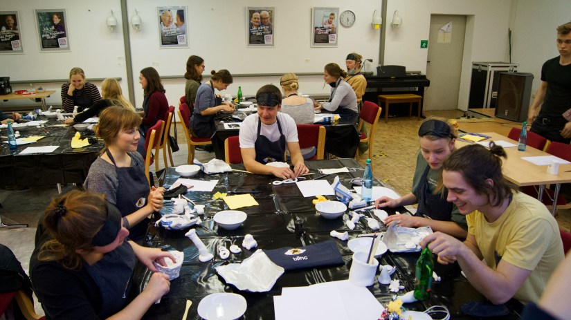 Zu sehen sind mehrere Tische mit  je 4 bis 5 Workshopteilnehmern. Diese machen sich mit dem Tonmaterial vertraut, zunächst noch ohne Augenmasken.