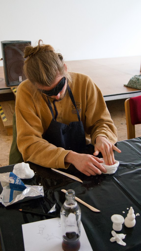 Eine Teilnehmerin mit schwarzer Augenmaske und Schürze sitzt am Tisch. Sie hält mit einer Hand die Tonschale fest und formt diese mit der anderen Hand.