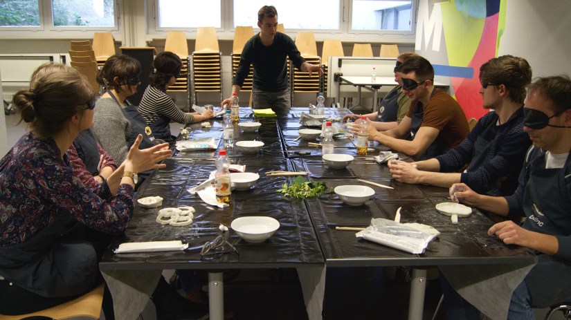 Ein Bild von einem Blinc Workshop mit unserem Workshopleiter Samuel und 6 Teilnehmern. Samuel steht vorne und erklärt etwas. Die Teilnehmer haben Augenmasken auf und formen ihre Tonkunstwerke. 