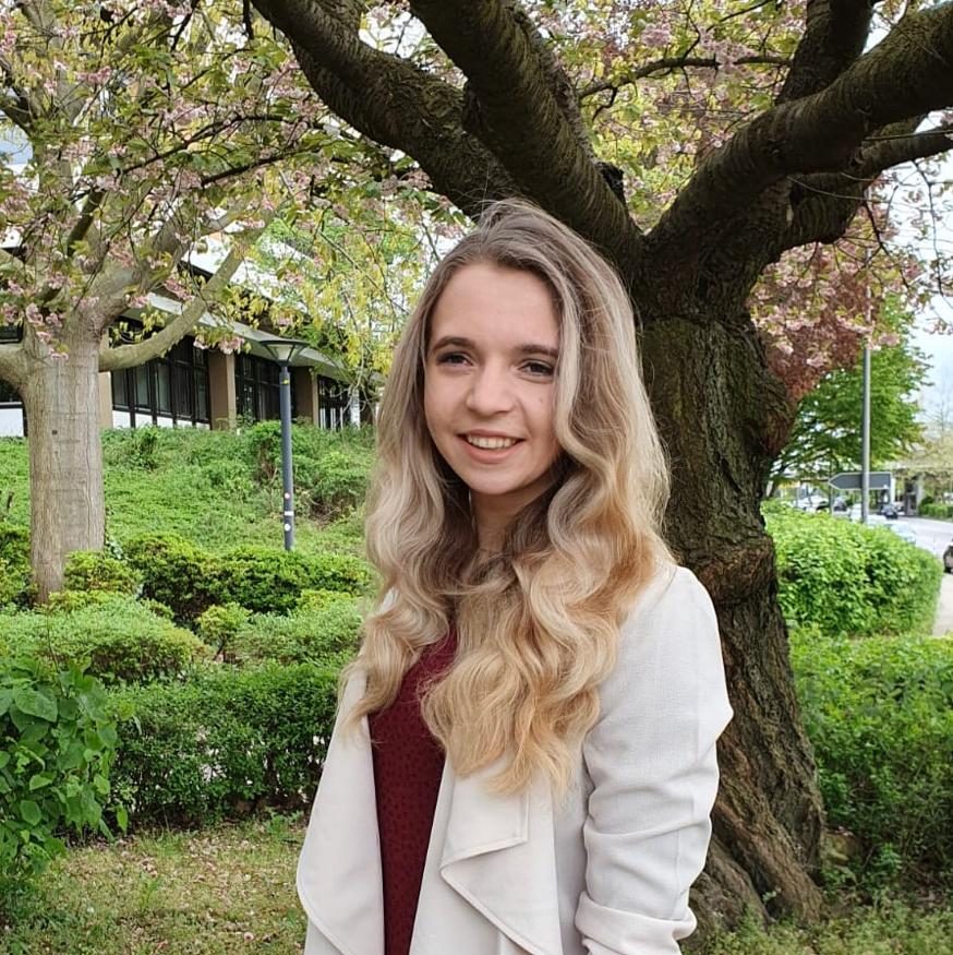 Hier siehst du unsere Workshopleiterin Vivien. Sie hat lange, wellig blonde Haare und lächelt in die Kamera. Hier trägt sie ein dunkelrotes Oberteil mit einem beigen Blazer mit Wasserfall Kragen.