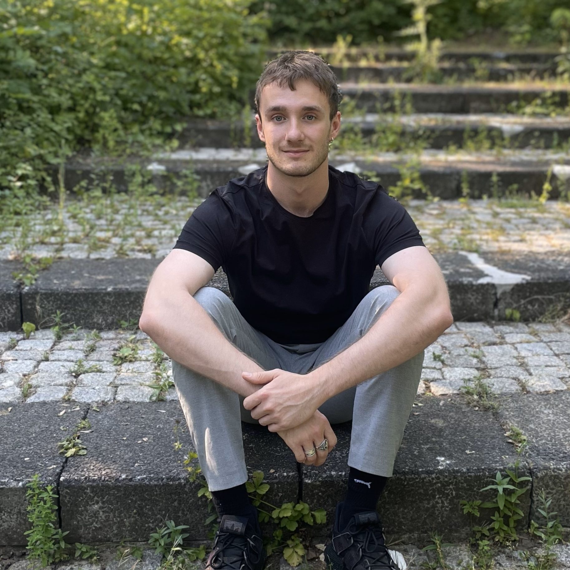 Hier sieht man Dominic. Auch er sitzt auf der Treppe im Schatten. Er hat kurze braune Haare und trägt ein schwarzes Tshirt und eine graue lange Hose. 