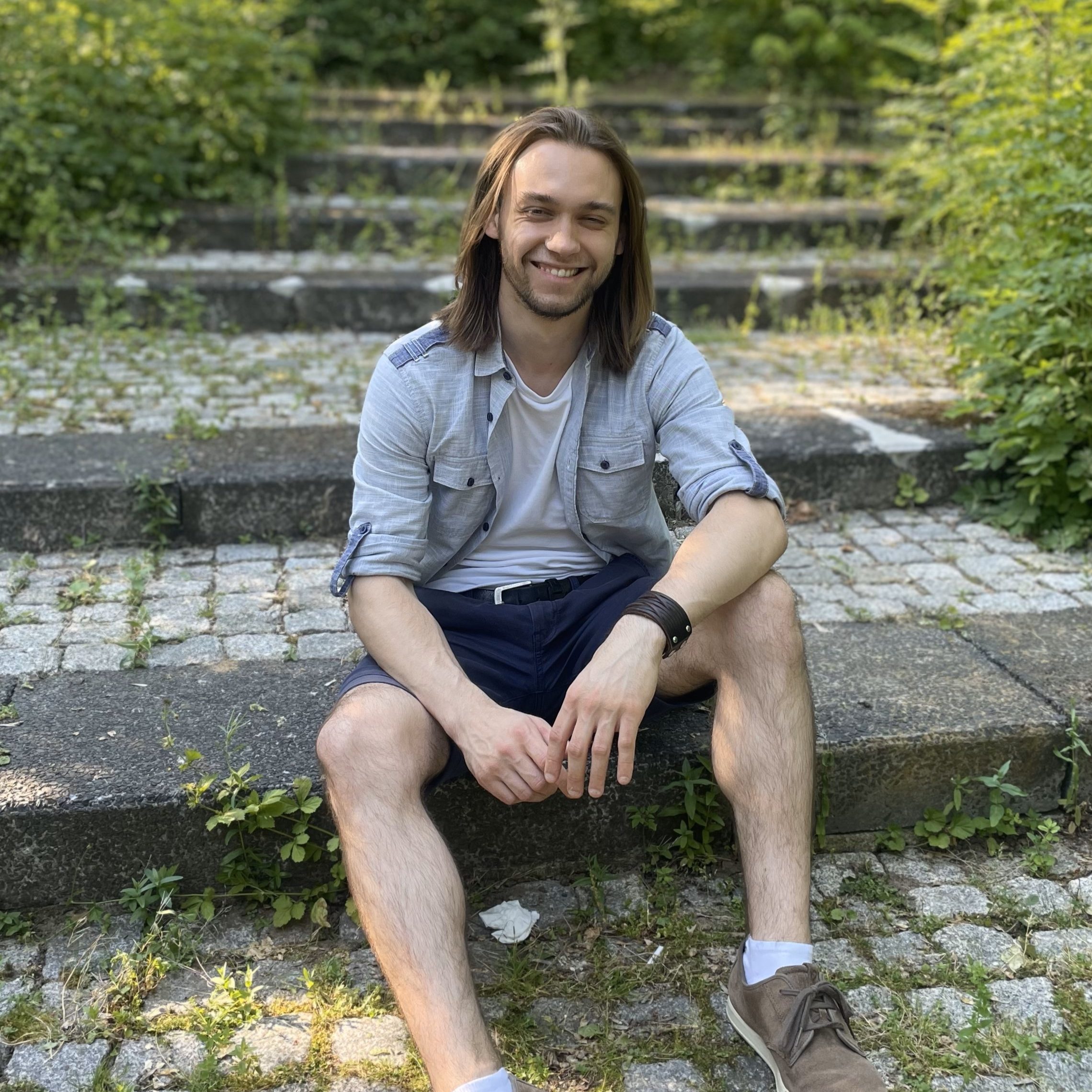 Hier sitzt unser Teamleiter Samu. Er hat braune, schulterlange Haare und lacht in die Kamera. Er trägt ein blaues Hemd über einem weißen Tshirt und eine kurze schwarze Hose.