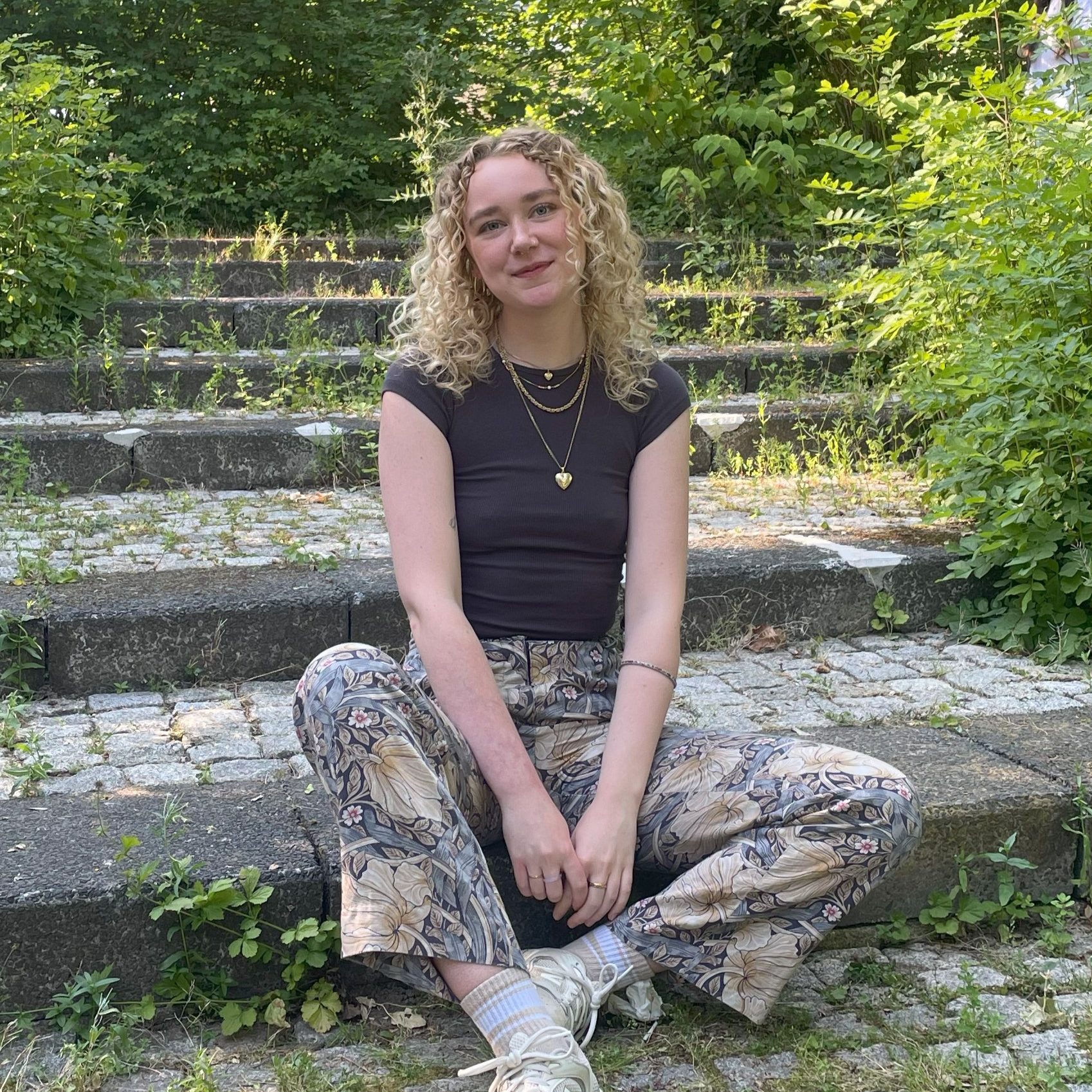 Hier sitzt die Teamleitung Marit auf einer Treppe. Sie hat lockige blonde Haare, lächelt und trägt ein schwarzes Tshirt. Zudem trägt sie eine lange Hose mit Blumenmuster.