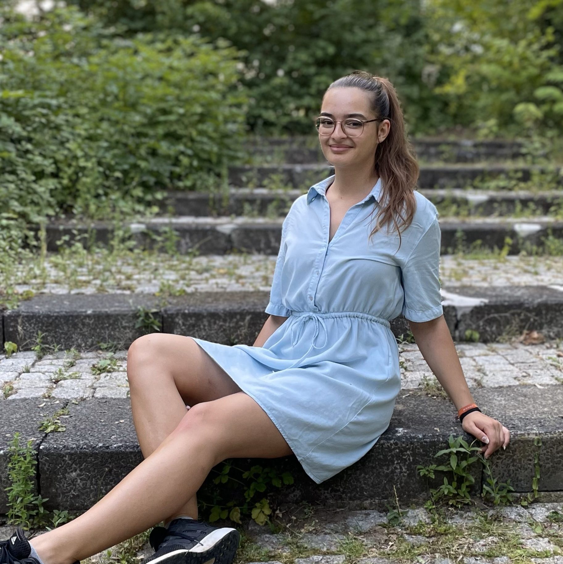 Hier sieht man Lia. Sie sitzt auf der Treppe und hat lange braune Haare, die zu einem Pferdeschwanz zusammengebunden sind. Sie trägt eine rosane Brille und ein hellblaues Hemdkleid.