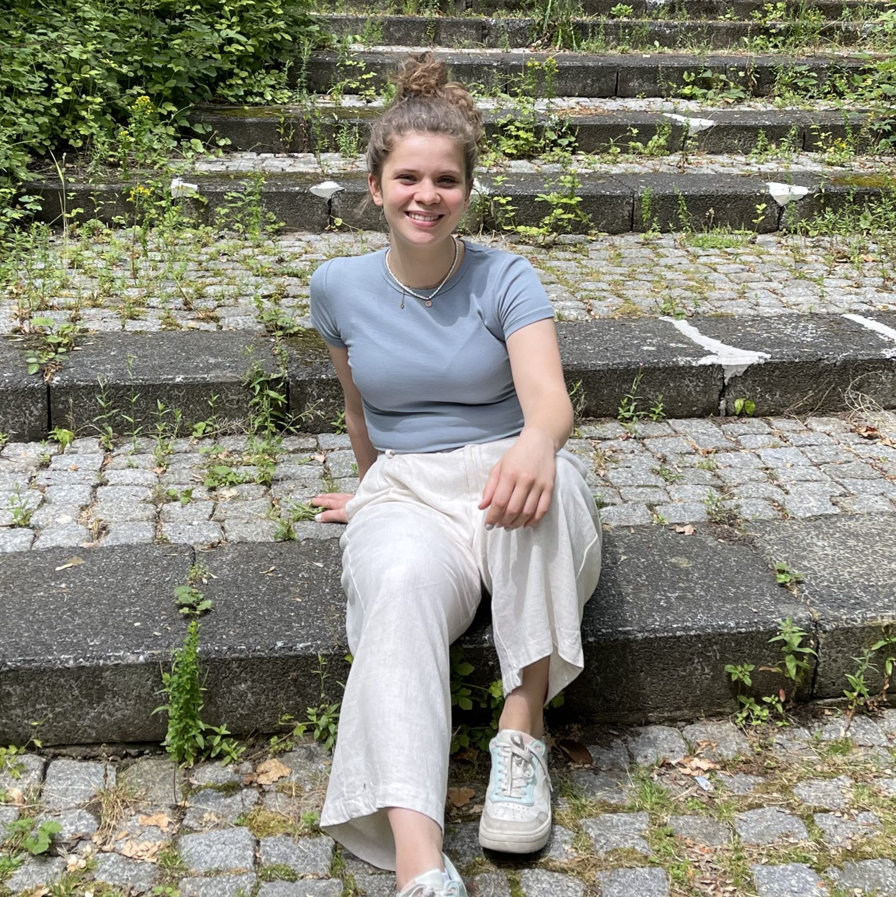 Hier sieht man Aylin. Sie sitzt in der Sonne und lächelt. Sie hat ihre lockigen Haare zu einem Dutt gebunden un dträgt ein hellblaues Tshirt und eine beige lange Hose. 